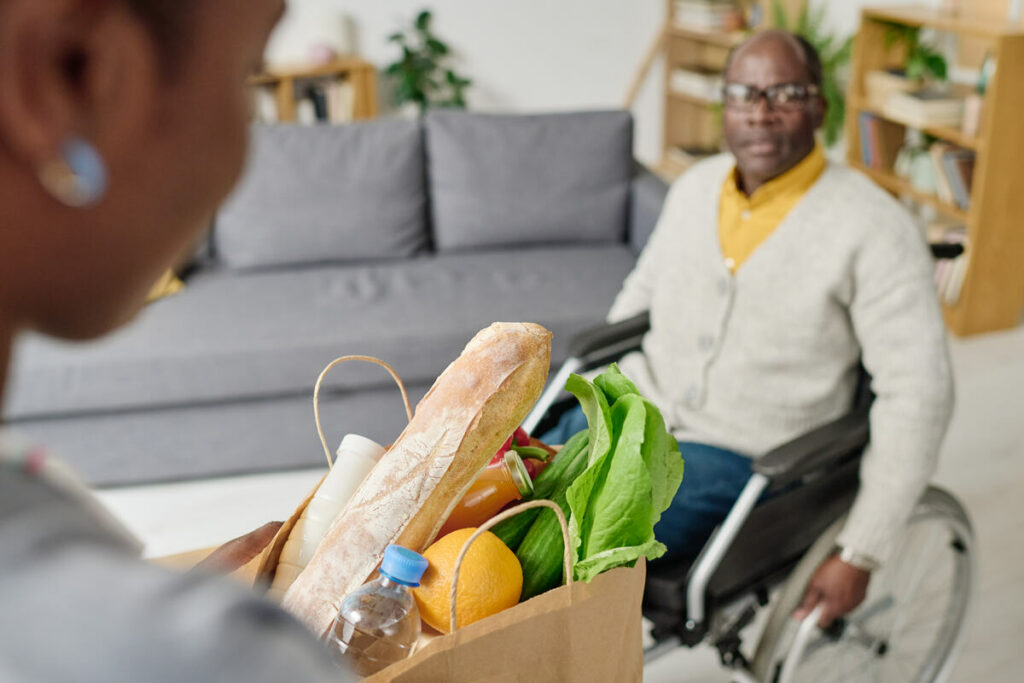 A person in a wheelchair receives a grocery bag with fresh produce from another person in a cozy living room.