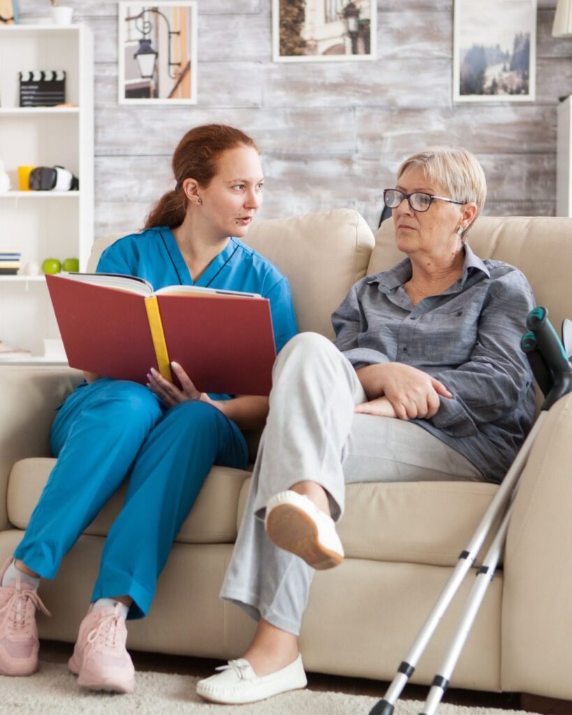 A caregiver reading a book with an elderly person, offering companionship and support in a comfortable home setting.