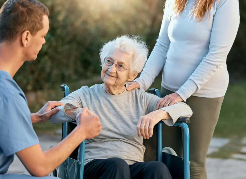 A caregiver assisting an elderly person in a wheelchair, with support from a family member, promoting mobility and personalized care in an outdoor setting.