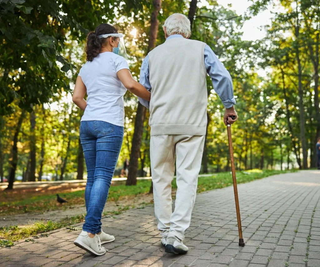 A caregiver assisting an elderly person with walking using a cane, promoting mobility and support during a peaceful walk in a park.