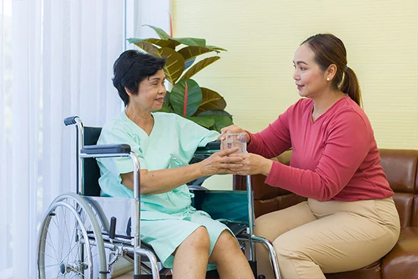 A caregiver assisting an elderly person in a wheelchair by providing a glass of water, promoting independence and support in a comfortable home setting.