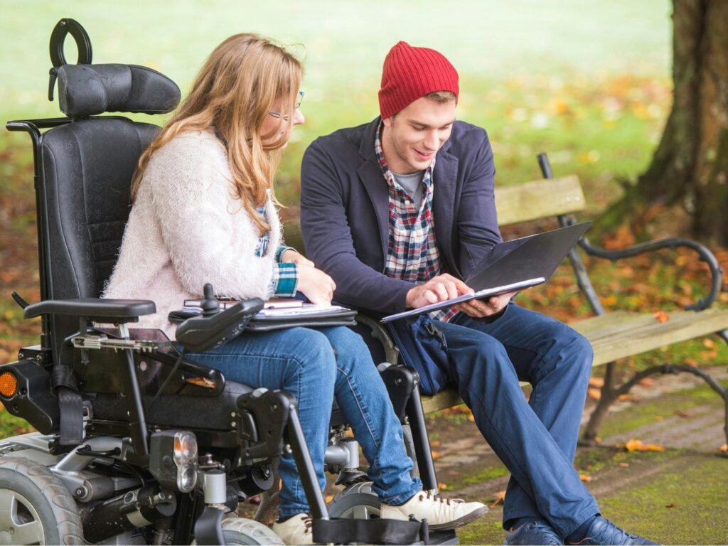 Two individuals, one in a wheelchair, sitting together on a park bench, looking at a laptop screen, enjoying the outdoors.