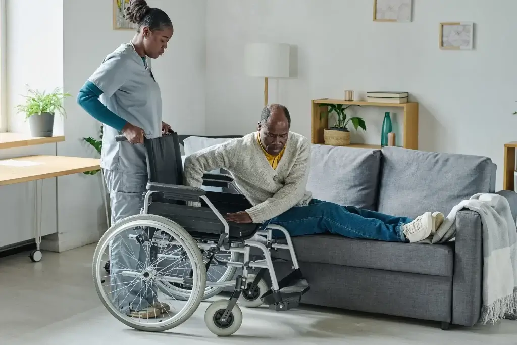 Caregiver assisting a person in a wheelchair to sit on a sofa in a comfortable home environment.