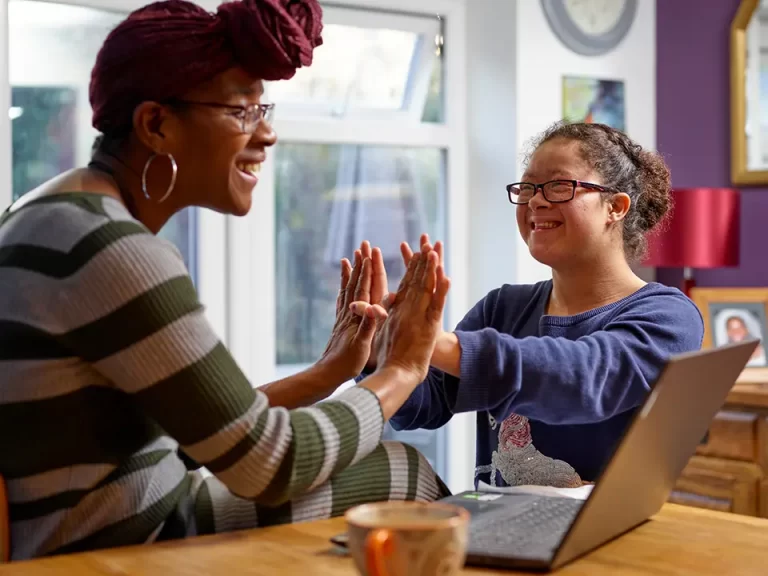 A caregiver and a young woman with glasses happily engaging in a hand game, promoting positive interaction and daily living skills in a home environment.