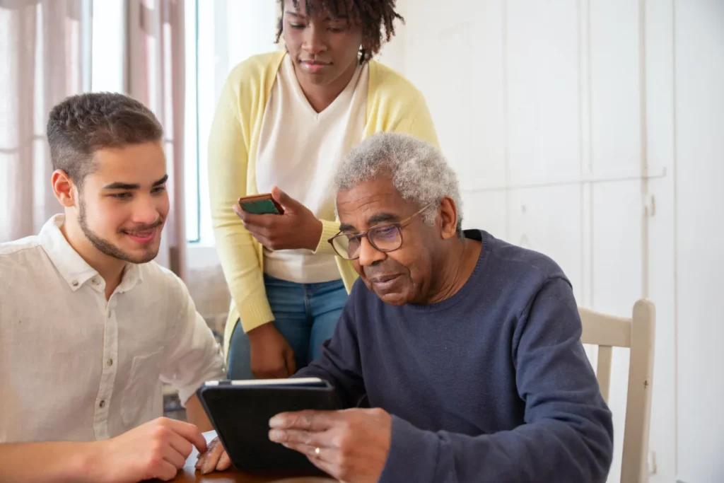 A caregiver assisting an elderly person with a tablet while two other individuals look on, promoting learning, technology use, and engagement in a supportive home environment.