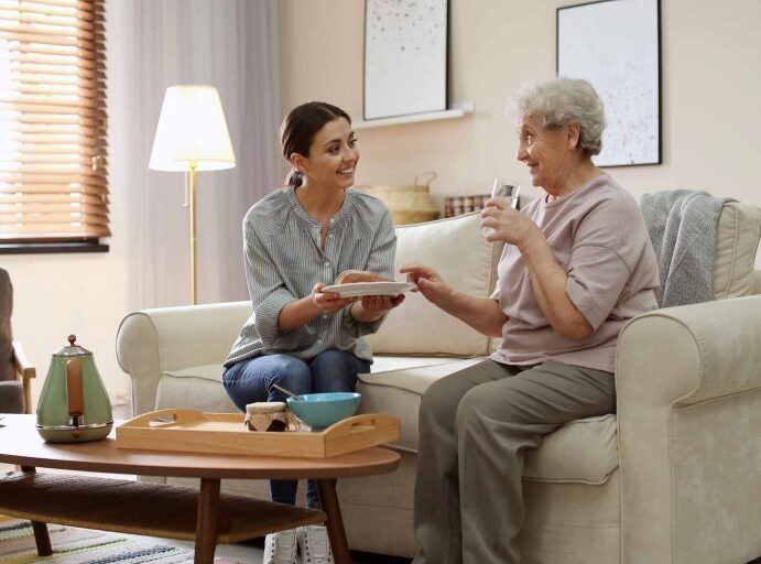 A caregiver serving food and offering a drink to an elderly person, promoting comfort and support in a cozy living room environment.